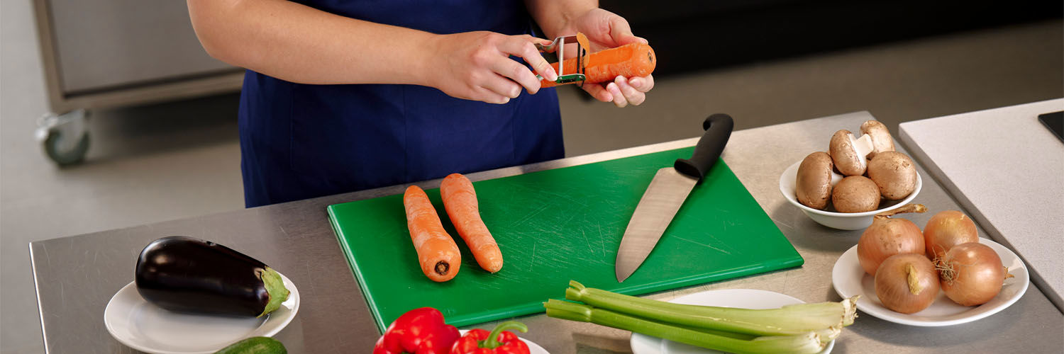 A person peels a carrot over a food preparation table laid with various vegetables.