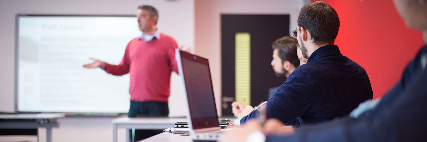 Lecturer Richard Bent delivering a tutorial session, he stands at the front of the class before the whiteboard while students at desks listen on.