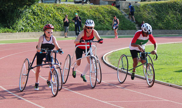 Three people using a three wheeled frame with a saddle, handlebars and a chest support (but no pedals) around a racetrack.