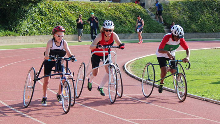 Three people using a three wheeled frame with a saddle, handlebars and a chest support (but no pedals) around a racetrack.