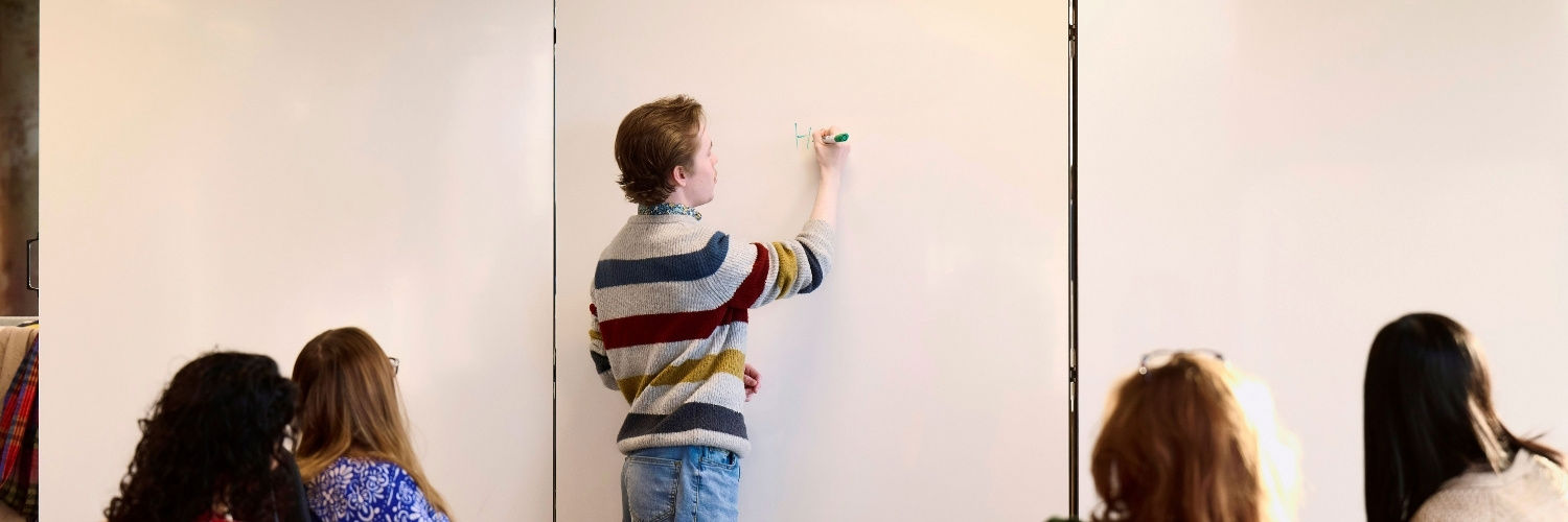 Male student writing on a white board while other students watch.