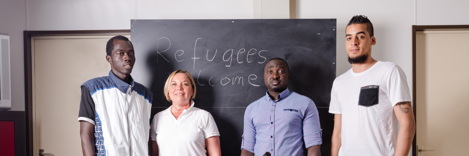 Image shows four men of colour and a white woman in a community hall in front of a chalkboard that reads "refugees welcome"