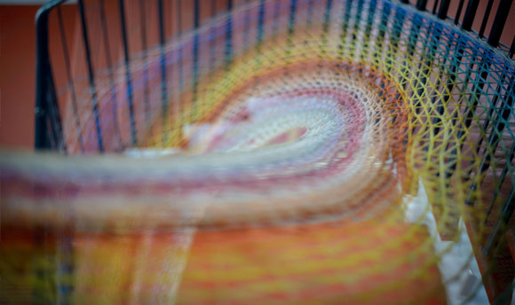 A close-up shot of a rainbow coloured piece of yarn work. The yarn appears to form a curved pattern suspended in the air.
