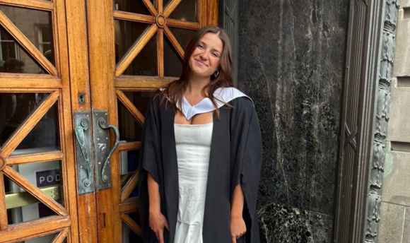 head and shoulders shot of female student with long brown hair in graduation robes and holding up certificate
