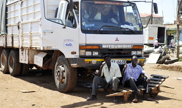 A white Mitsubishi flatbed truck parked on a dirt ground, two individuals sit on a bench in front that appears to be fashioned from wooden pallets.