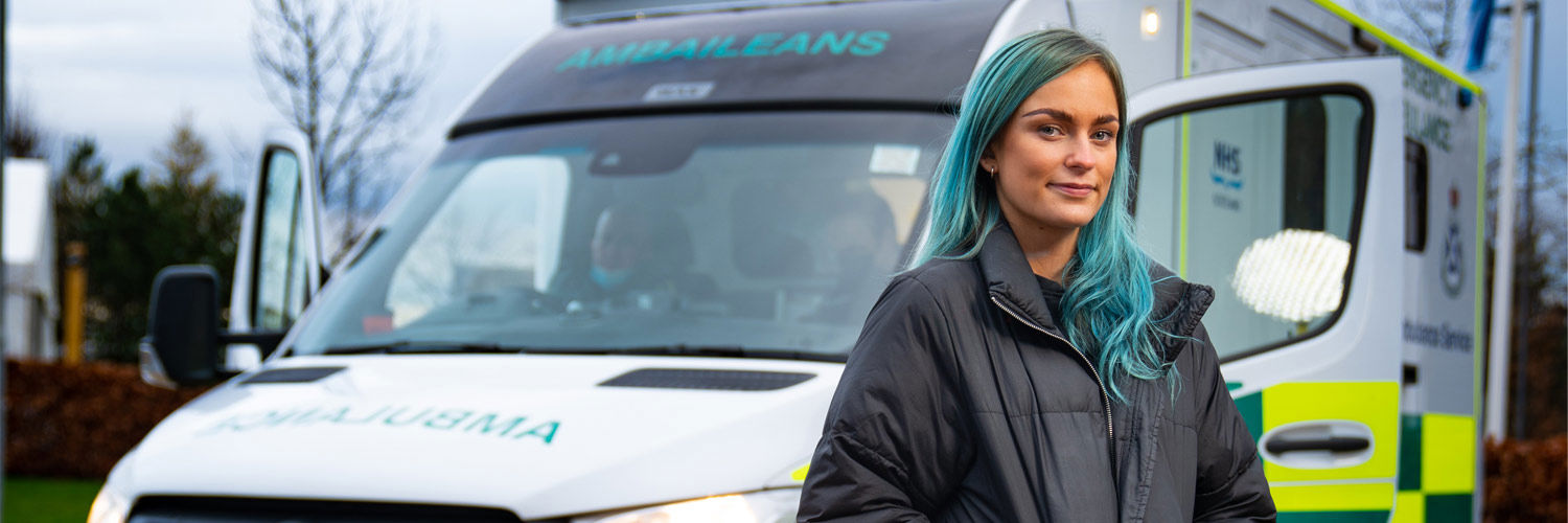 A student with teal-coloured hair stands in front of an ambulance.