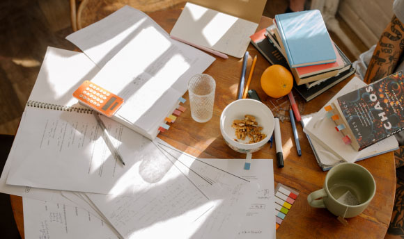 A table scattered with different books, notepads, stationery and snacks.