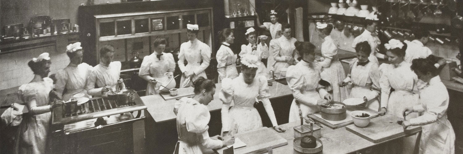 B+W photo of a kitchen full of women in Edwardian style chef's whites
