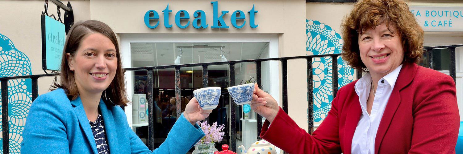 Two women smiling, holding up china tea cups outside a cafe called "eteaket"