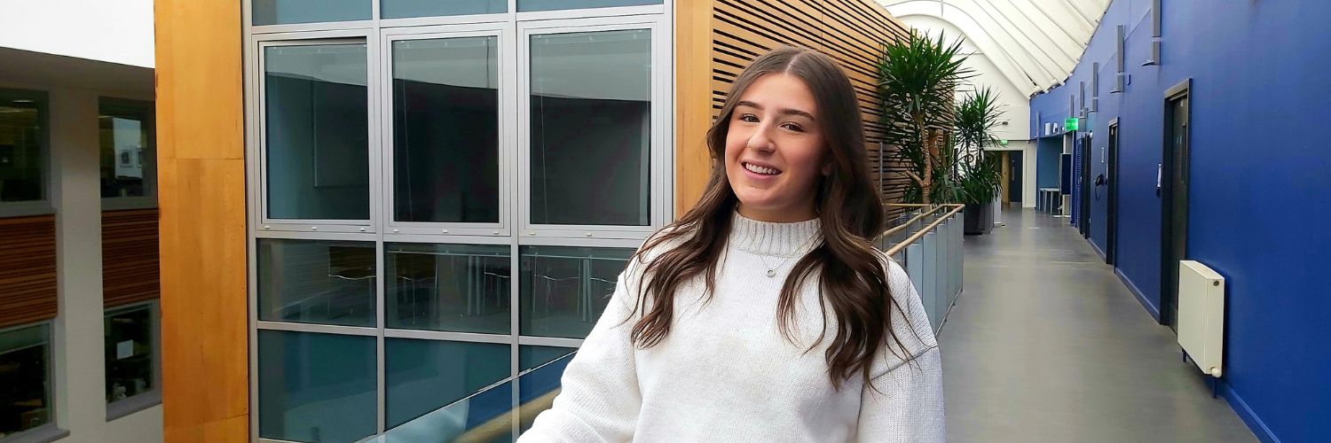 Young female student with long brown hair with backdrop of the interior of Queen Margaret University 