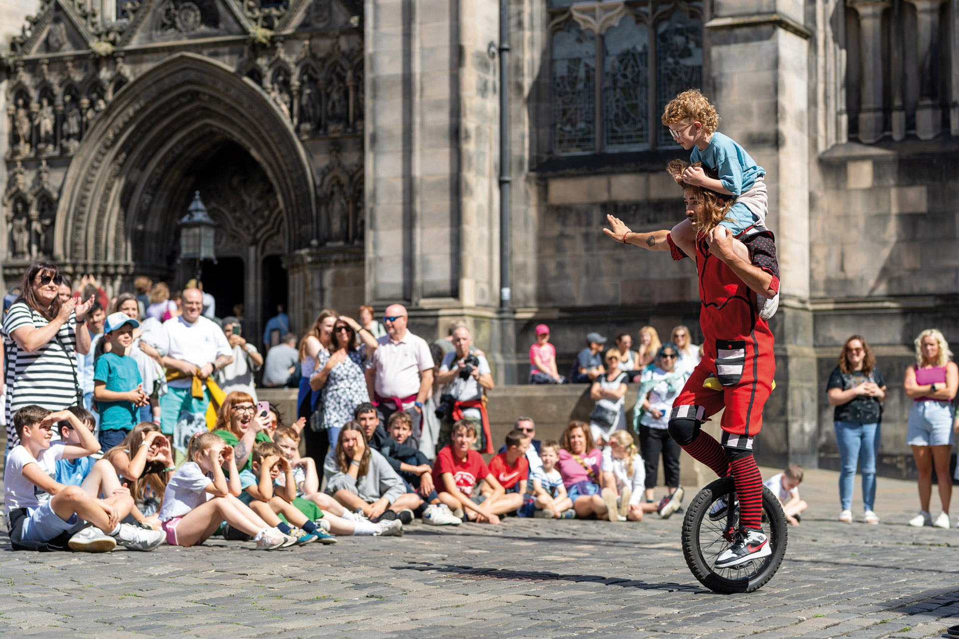 Image of Edinburgh Festival Street Performer on a Unicycle with a you child on his shoulder in front of a crowd