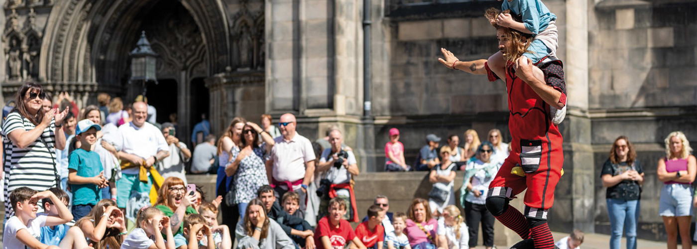 Image of Edinburgh Festival Street Performer on a Unicycle with a you child on his shoulder in front of a crowd
