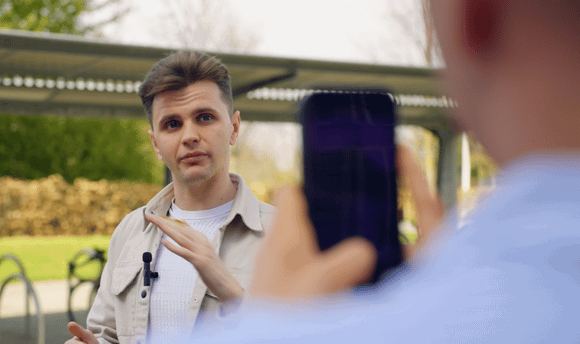 A male student speaking and being filmed by another student on a phone.