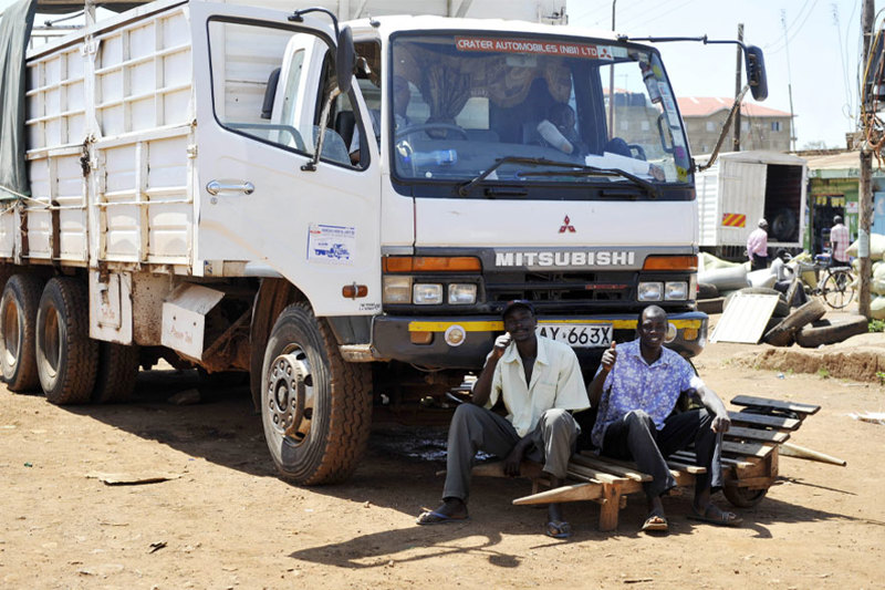 A white Mitsubishi flatbed truck parked on a dirt ground, two individuals sit on a bench in front that appears to be fashioned from wooden pallets.