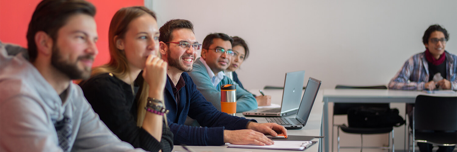 Queen Margaret University International Management with Leadership students sitting in class smiling with laptops or pads of paper and pens infront of them