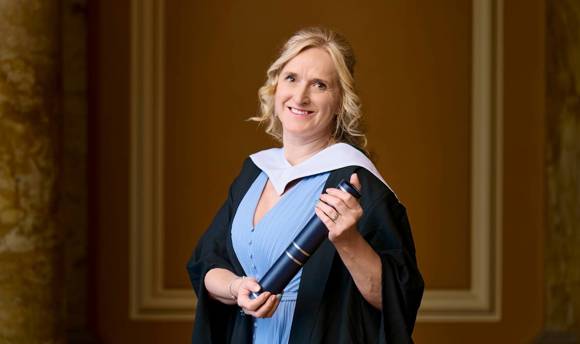 portrait style image of female blonde student wearing graduation robe and holding graduation scroll 