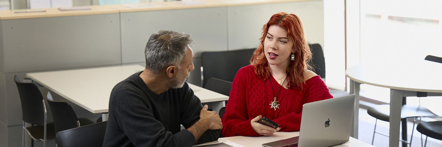 Two people seated, chatting together beside a laptop device.