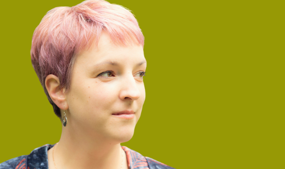 Headshot of a white woman with short cropped pink hair and earrings posing against a green background.