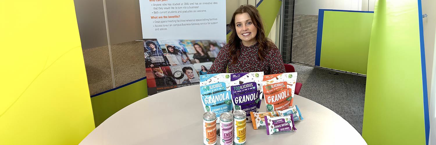 girl sitting behind table covered in colourful cans of energy drinks and packets of granola