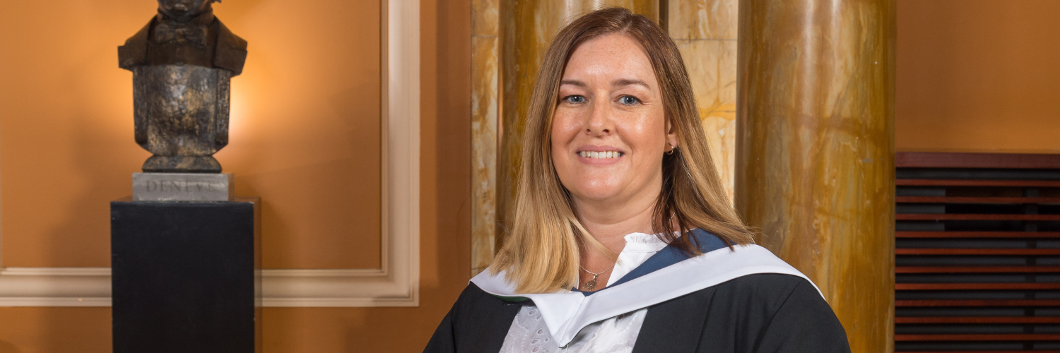 Close up image of a woman with blonde shoulder length hair smiling wearing her graduation robe and holding her graduation scroll
