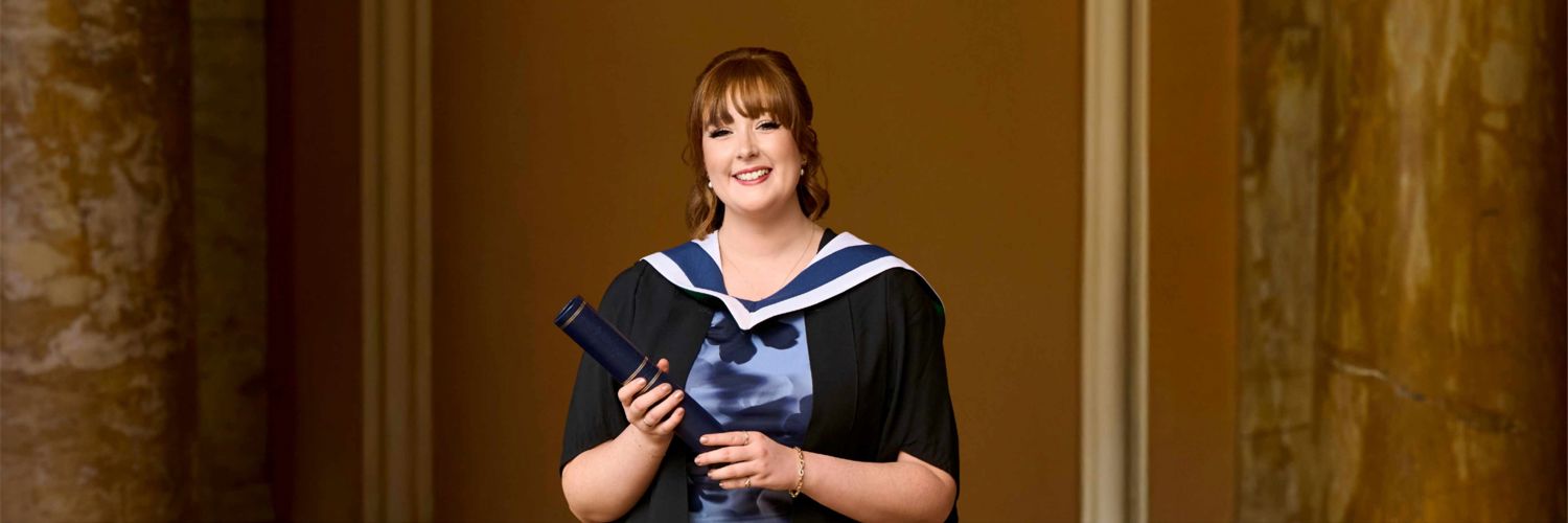 Head and shoulders shot of smiling student wearing graduation robes and holding graduation scroll