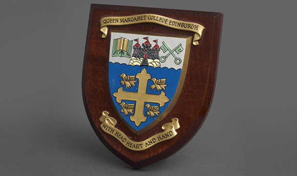 The Queen Margaret University College crest and motto, "with Head, Heart and Hand" affixed to a wooden shield.