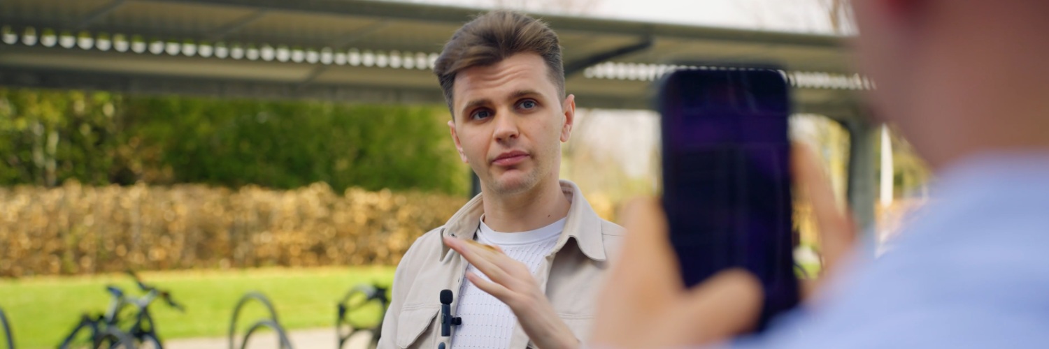 A male student speaking and being filmed by another student on a phone.