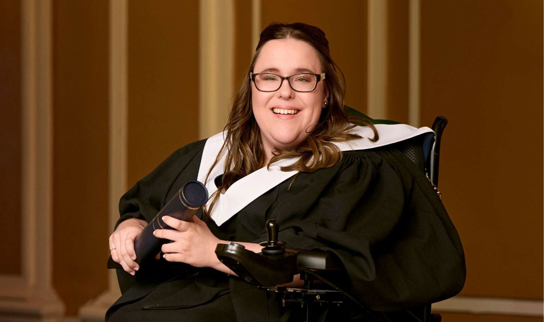 portrait style image of Charlotte sitting in wheelchair  wearing graduation robe and holding graduation scroll 