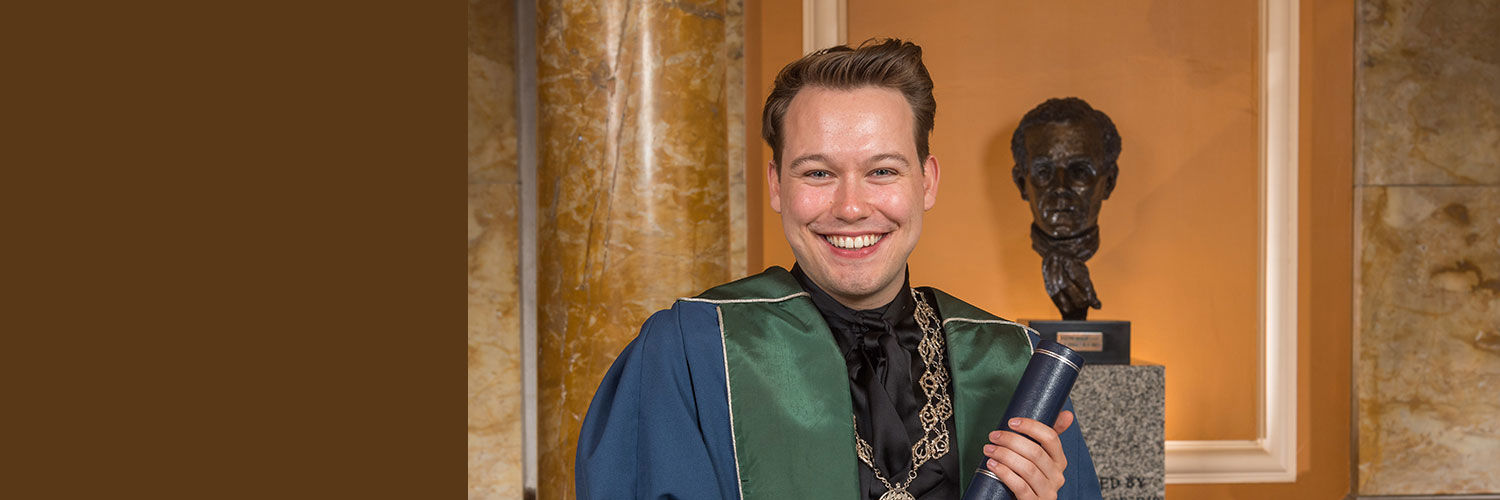 Portrait image of Jay Brown wearing his graduation robes and holding a graduation scroll