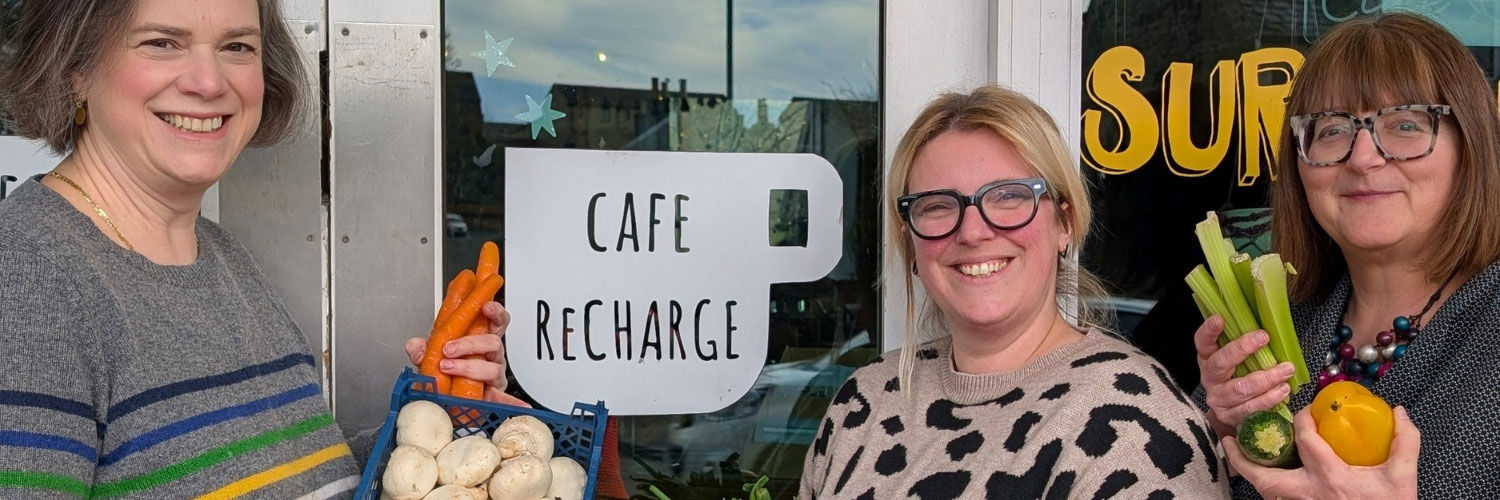 Three people pose for a photo outside of a cafe, holding an assortment of vegetables.