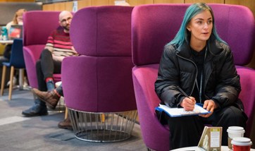 Student with vibrant blue hair sat on purple pod seat in QMU Atrium