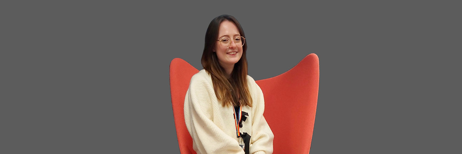 A young female student with brown hair and glasses sits in a bright orange-red chair.