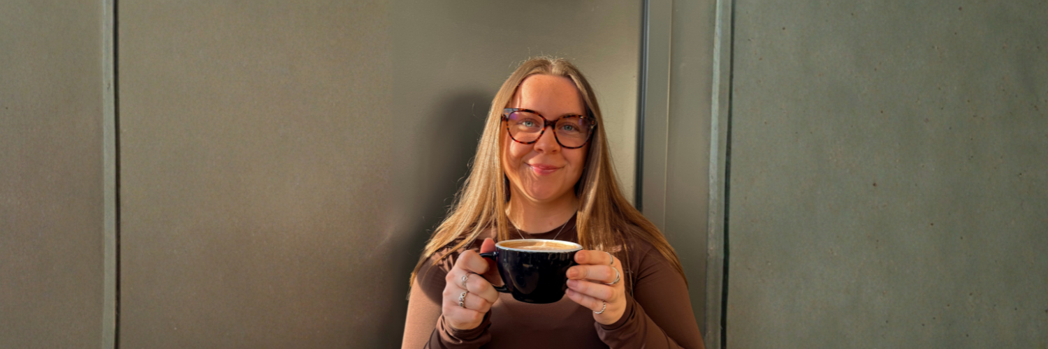 QMU Student Ellie holding a mug of coffee and smiling for the camera. 