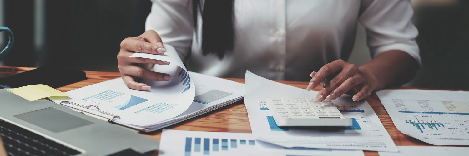 A woman using a calculator with financial reports surrounding her on a desk. 