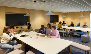 A teaching space within the library, there are two large tv screens, a whiteboard, and two long tables filled with students working on laptops.