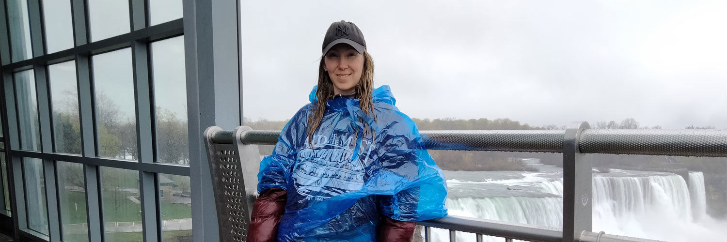 A young woman wearing a hat and poncho stands in front of Niagara falls