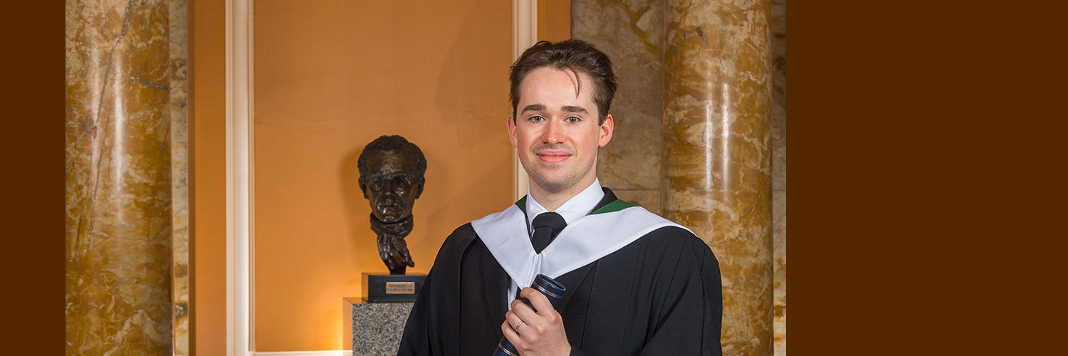Portrait shot of QMU graduate Calan dressed in black graduation robes holding graduation scroll