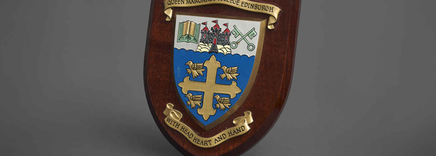 The Queen Margaret University College crest and motto, "with Head, Heart and Hand" affixed to a wooden shield.