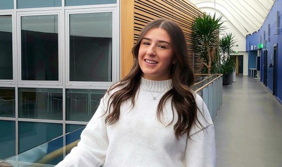 Young female student with long brown hair with backdrop of the interior of Queen Margaret University 