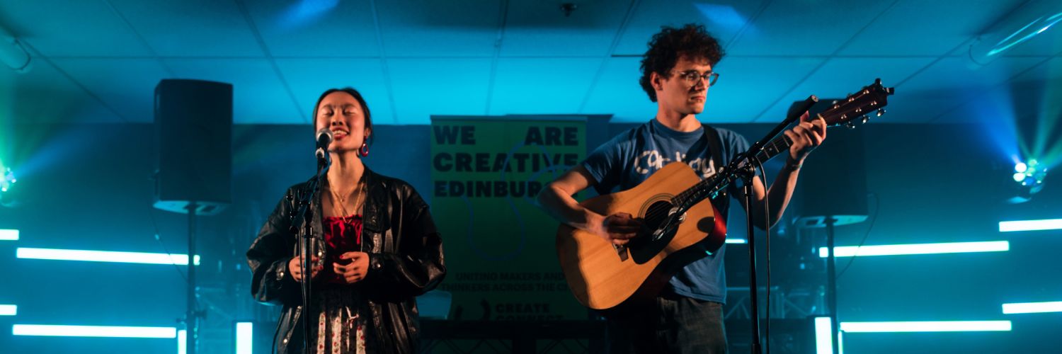 A young woman and a young man performing a song in a dark venue with a Creative Edinburgh sign behind them.