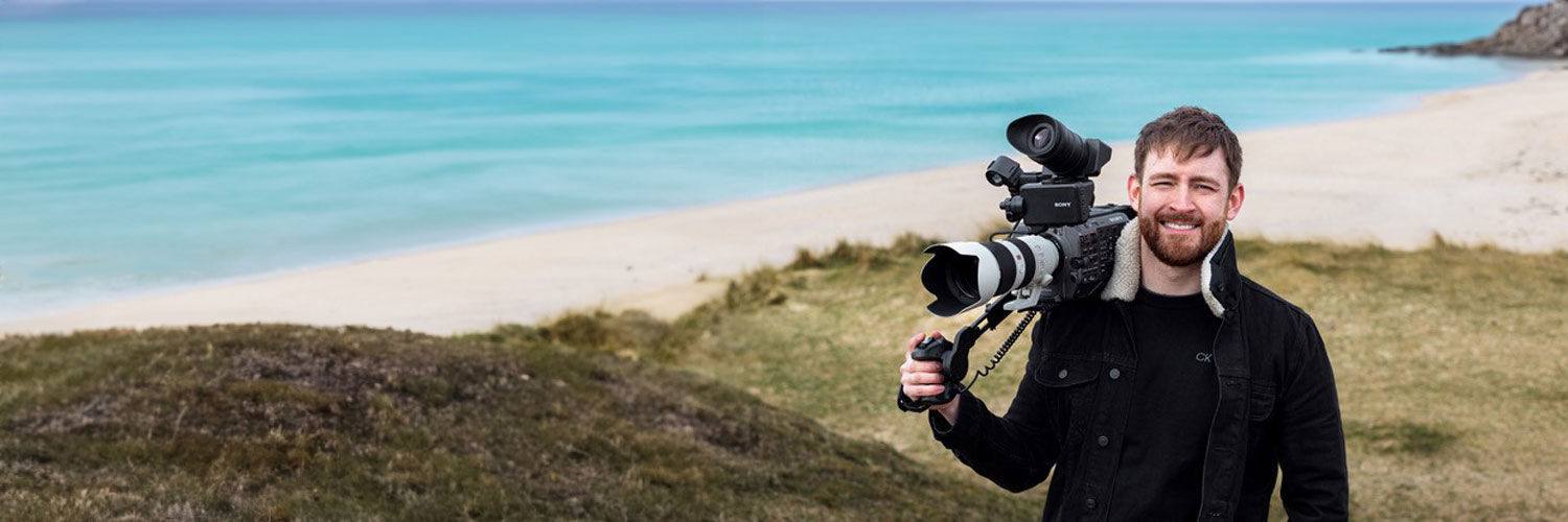 A young man holding aloft a state of the art video camera poses on a grassy hill facing down onto the seaside.