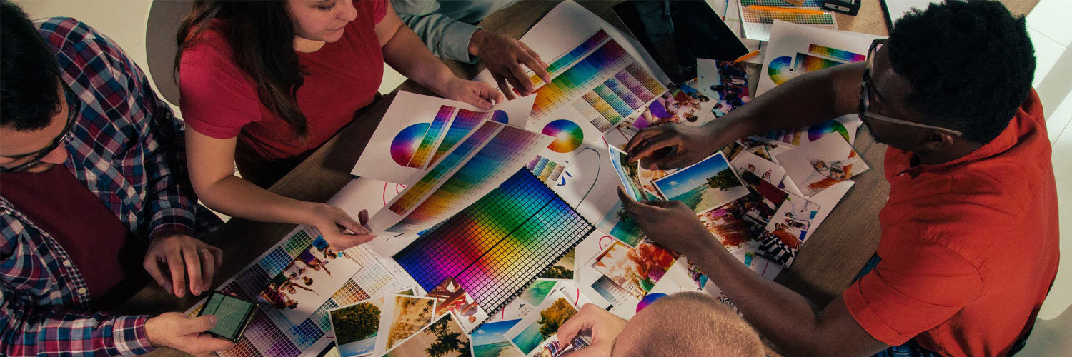 A group of four students sat at a desk together. They are browsing through a variety of different print materials featuring colour gradients and professionally-taken photographs. [Credit: iStock]