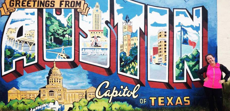A person poses next to a stylised billboard that reads "Greetings from Austin, Capital of Texas".