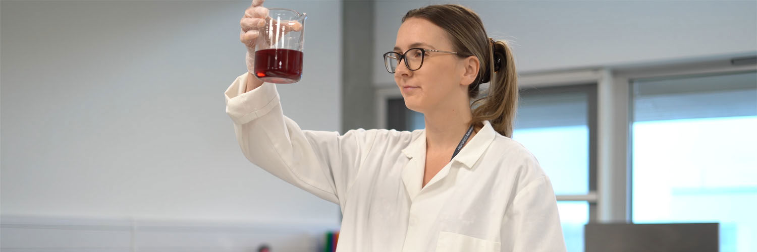 A woman wearing a lab coat, holding up a beaker of liquid.