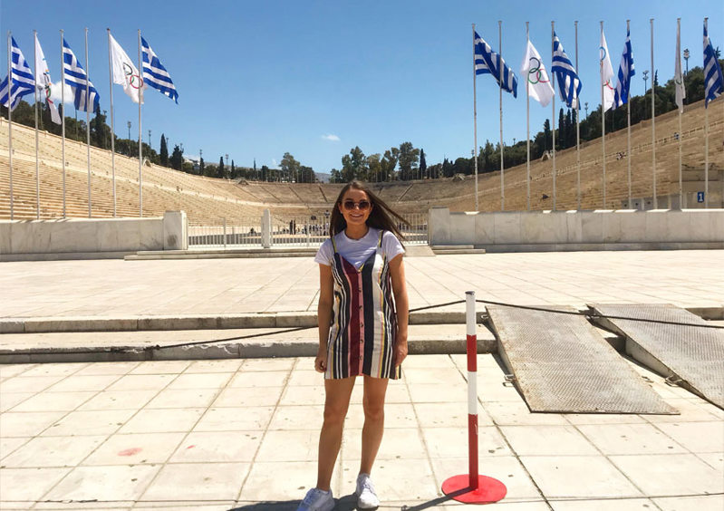 A person poses for a photo, various Greek and Olympics flags stand on tall flagpoles behind them.