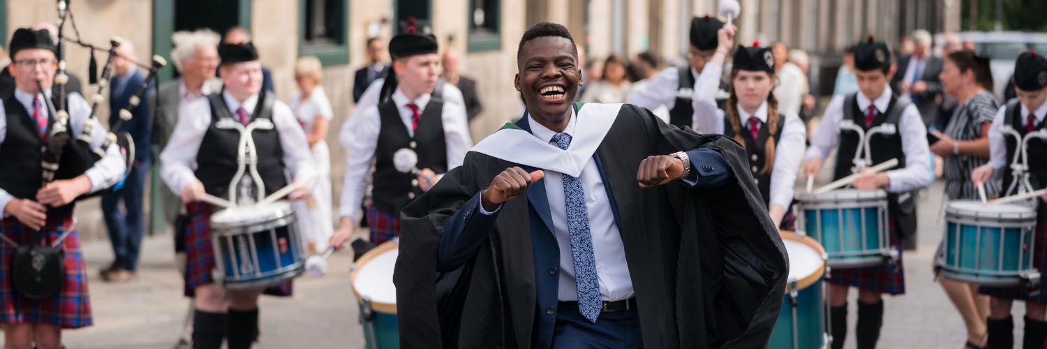 a young black man with a big smile, wearing his graduation gown and dancing in front of a pipe band playing outside the graduation venue