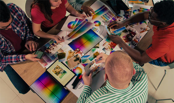 A group of four students sat at a desk together. They are browsing through a variety of different print materials featuring colour gradients and professionally-taken photographs. [Credit: iStock]
