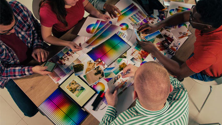 A group of four students sat at a desk together. They are browsing through a variety of different print materials featuring colour gradients and professionally-taken photographs. [Credit: iStock]
