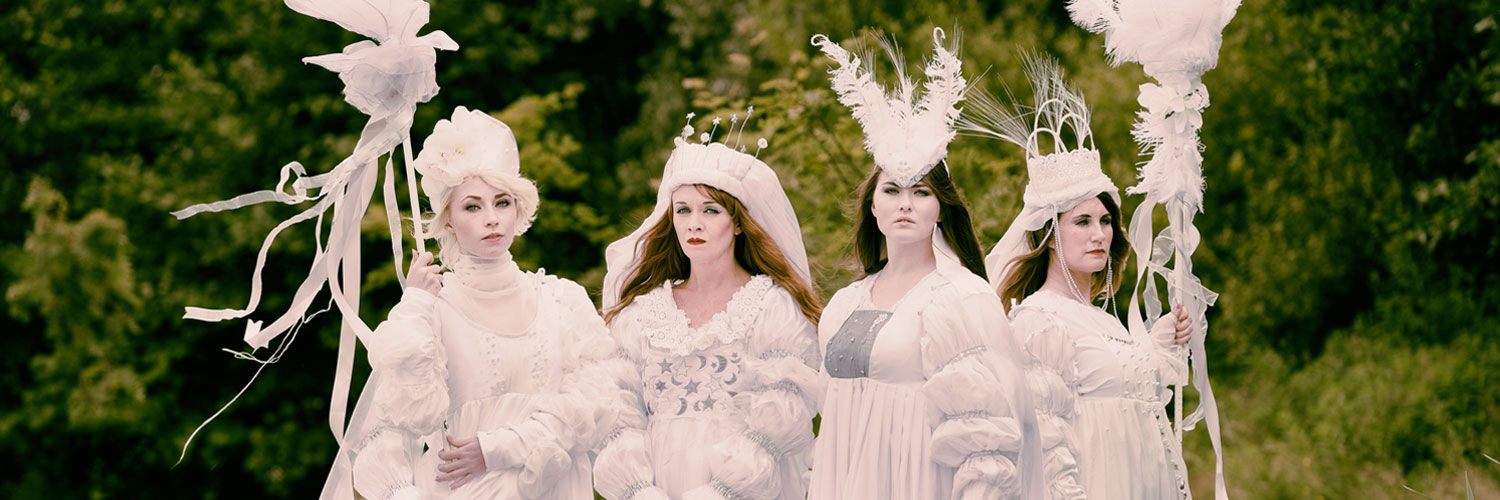 Four women wearing ornate, all-white robes, feathered hairpieces and holding white staffs. Their outfits are themed around 'Selene, goddess of the moon'.