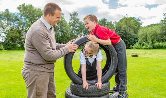 A small boy and girl playing with a tyre in a playpark a man holding the tyre so the girl can climb through the middle
